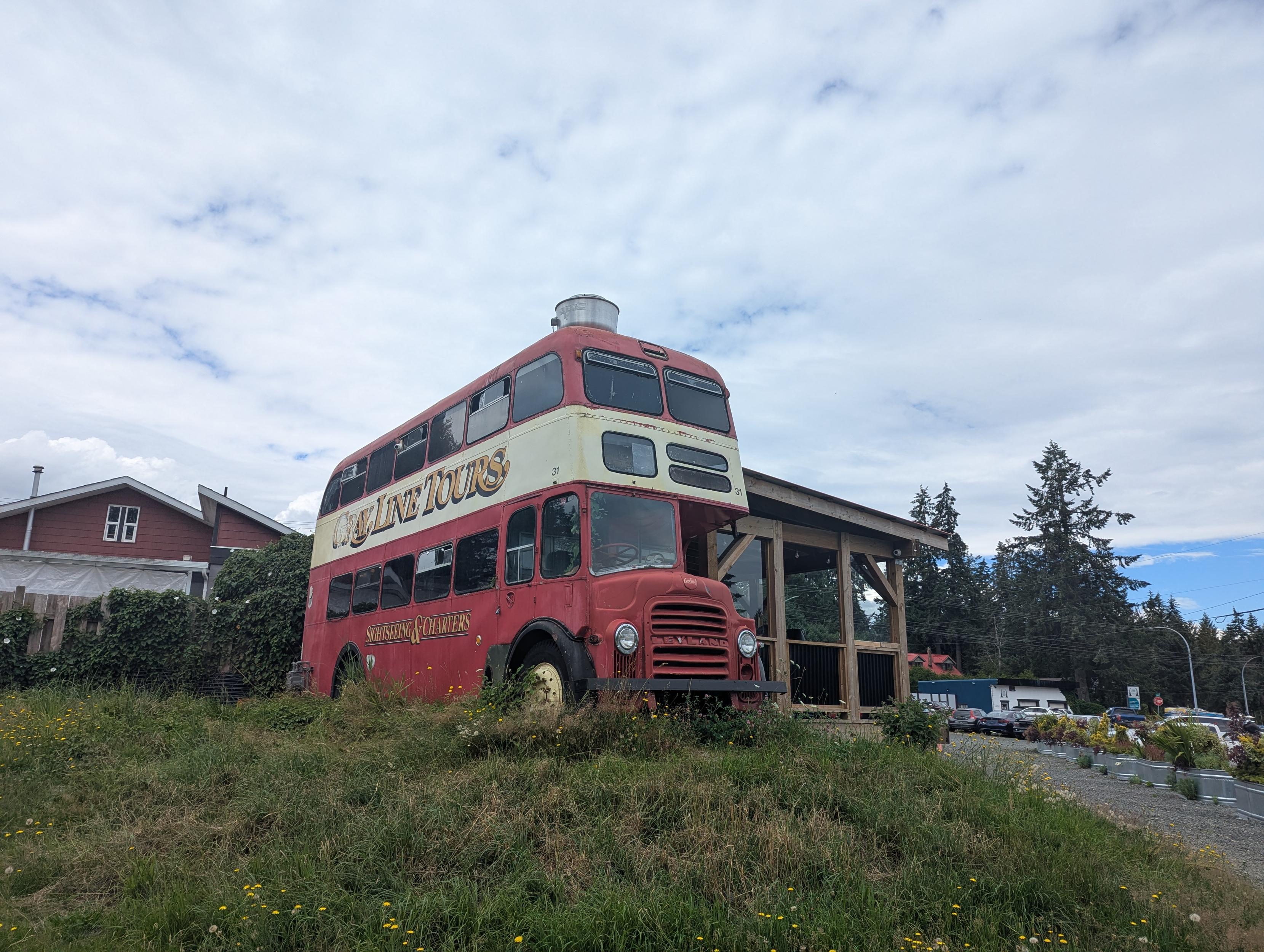An old-timey double decker bus with an attached shack on a grassy knoll. The bus is faded red and beige, "Grey line tours, sightseeing and charters". The sky is mostly white with bits of blue peeking out.