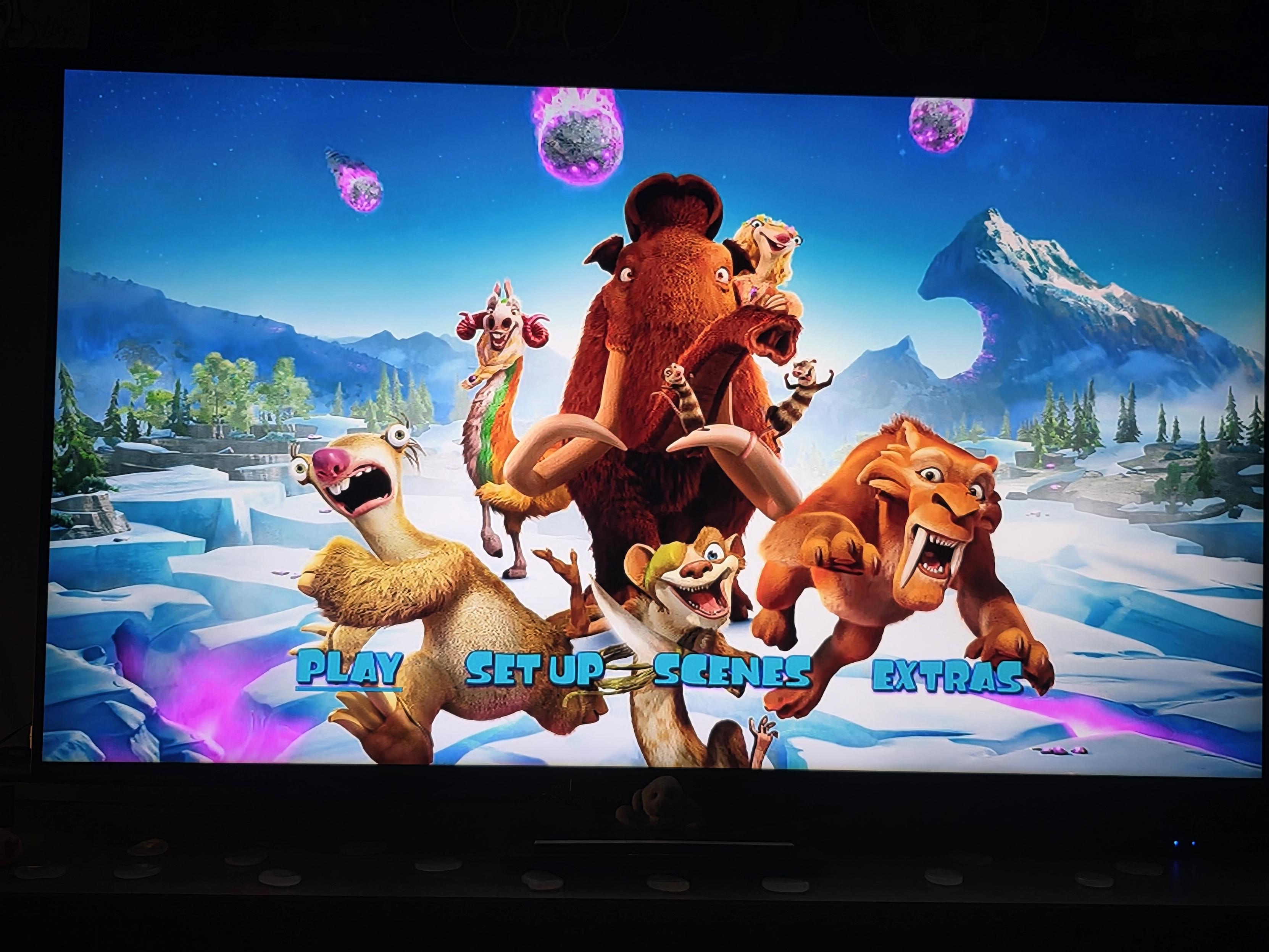 ​A photo of Cynni’s TV screen displaying the main DVD menu for Ice Age: Collision Course. The colorful, vibrant background shows a snowy prehistoric landscape under a starry blue sky filled with glowing purple and pink meteors falling from above.
​In the foreground, the main characters are posed dynamically together: Sid the sloth is on the left, looking goofy with wide eyes and his mouth open; Manny the woolly mammoth stands prominently in the center, looking surprised with his trunk raised; Diego the saber-toothed tiger is on the right, pouncing forward with a wild expression; Buck the eye-patched weasel smiles mischievously at the bottom; Shangri Llama stands colorful in the mid-ground; Brooke the sloth rests atop Manny’s back; and the possum brothers Crash and Eddie hang off Manny’s tusks.
​Near the bottom of the screen, bright cyan menu options with purple outlines are displayed over a glowing pink haze: "PLAY", "SET UP", "SCENES", and "EXTRAS". The bezel of the dark TV frame is visible around the screen.