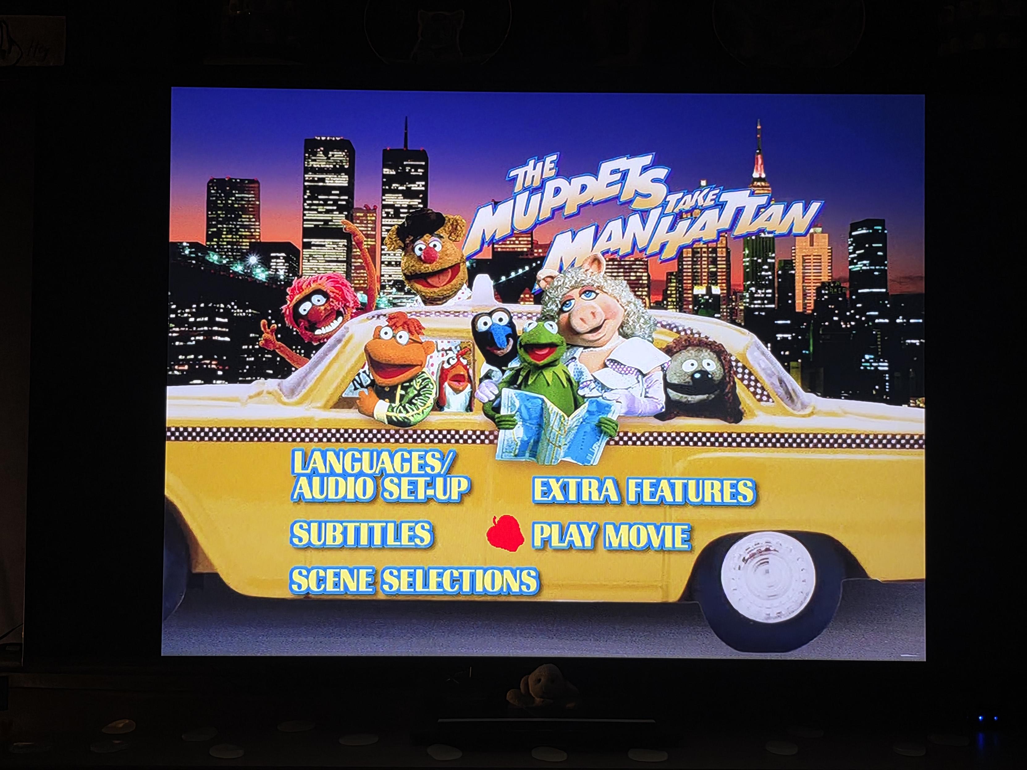 A photo of Cynni’s TV screen showing the main DVD menu for The Muppets Take Manhattan. In the background, the glowing New York City skyline is lit up against a deep purple and twilight sky.
​In the center of the frame, a classic yellow New York taxi cab is packed full of Muppets looking out the windows. Animal waves wildly from behind the cab; Fozzie Bear wears a driver's hat in the driver seat; Scooter leans out the side; Gonzo peeks out next to Kermit the Frog, who holds an open blue map; Miss Piggy sits elegantly in a white outfit and curly hair; and Rizzo the Rat rests on the far right. Above the taxi, the stylized film title "THE MUPPETS TAKE MANHATTAN" is written in bright blue and white 3D lettering.
​The yellow side of the taxi features the DVD menu options written in bold blue text: "LANGUAGES / AUDIO SET-UP", "SUBTITLES", "SCENE SELECTIONS", "EXTRA FEATURES", and "PLAY MOVIE", with a small red apple icon highlighting the play option. The black television frame borders the image.