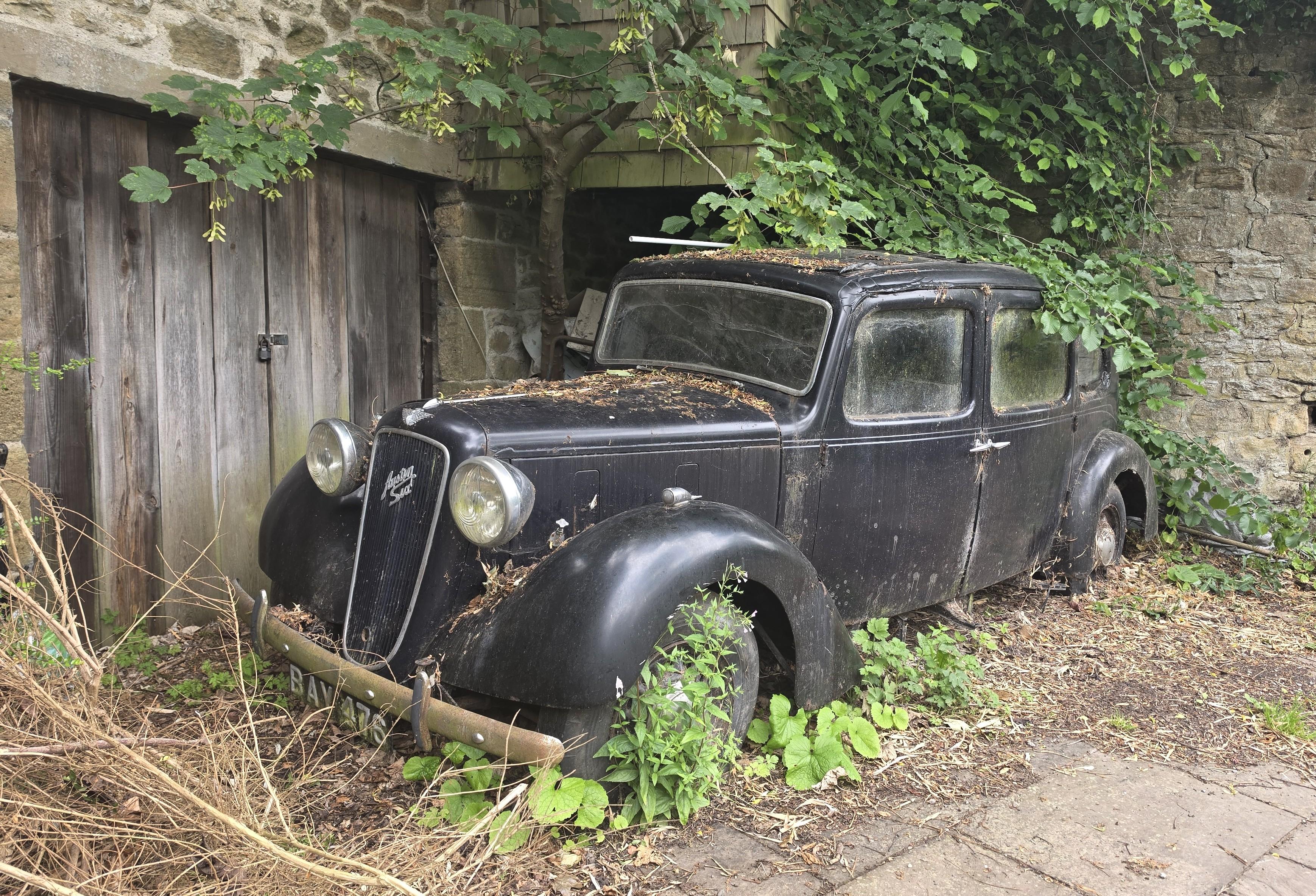 Picture of an abandoned black car, overgrown and sad looking. It's standing against the stone walls of a building with a large and rather dilapidated looking wooden door. It's an Austin and looks like it could be from the fourties or fifties, now resting near Corbridge, England.