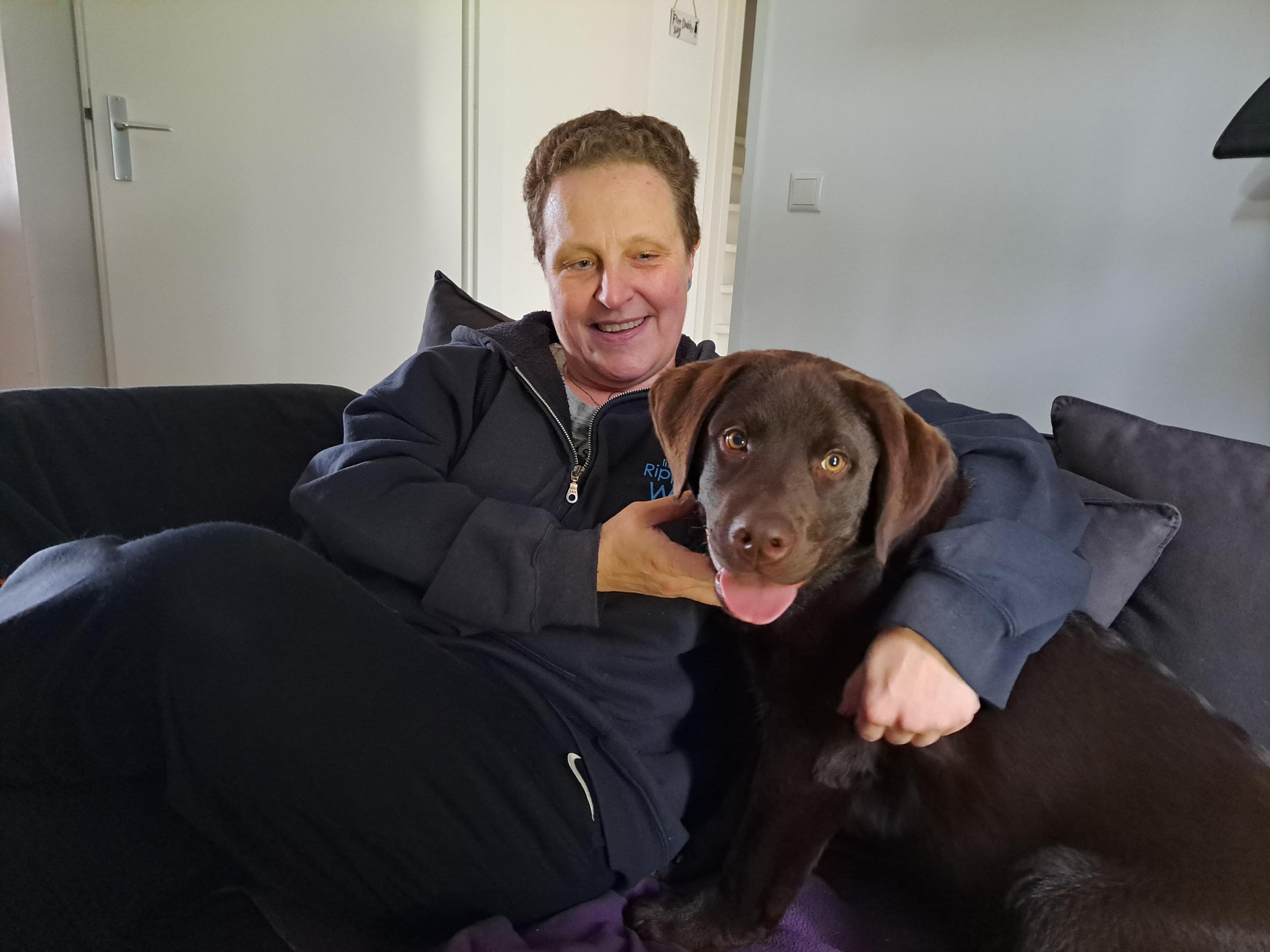 A warm and cozy indoor photo taken by Yolande, capturing a sweet moment between Cynni and their chocolate brown Labrador Retriever puppy, Koa. Cynni is sitting relaxed on a dark grey fabric couch, wearing a dark zip-up hoodie over a grey shirt and black pants. They have short brown hair and a gentle, affectionate smile as they look lovingly down at Koa.

Koa, a young puppy with smooth dark brown fur, bright amber-brown eyes, and floppy ears, sits attentively right beside Cynni. He is looking directly at the camera with a happy expression, his pink tongue playfully sticking out slightly. Cynni has one arm wrapped tenderly around Koa, holding him close in a warm embrace while gently resting their hand near his chin. The background shows a bright, softly lit room with a white door and light grey walls, highlighting the sweet bond and comfortable warmth shared between Cynni and Koa.