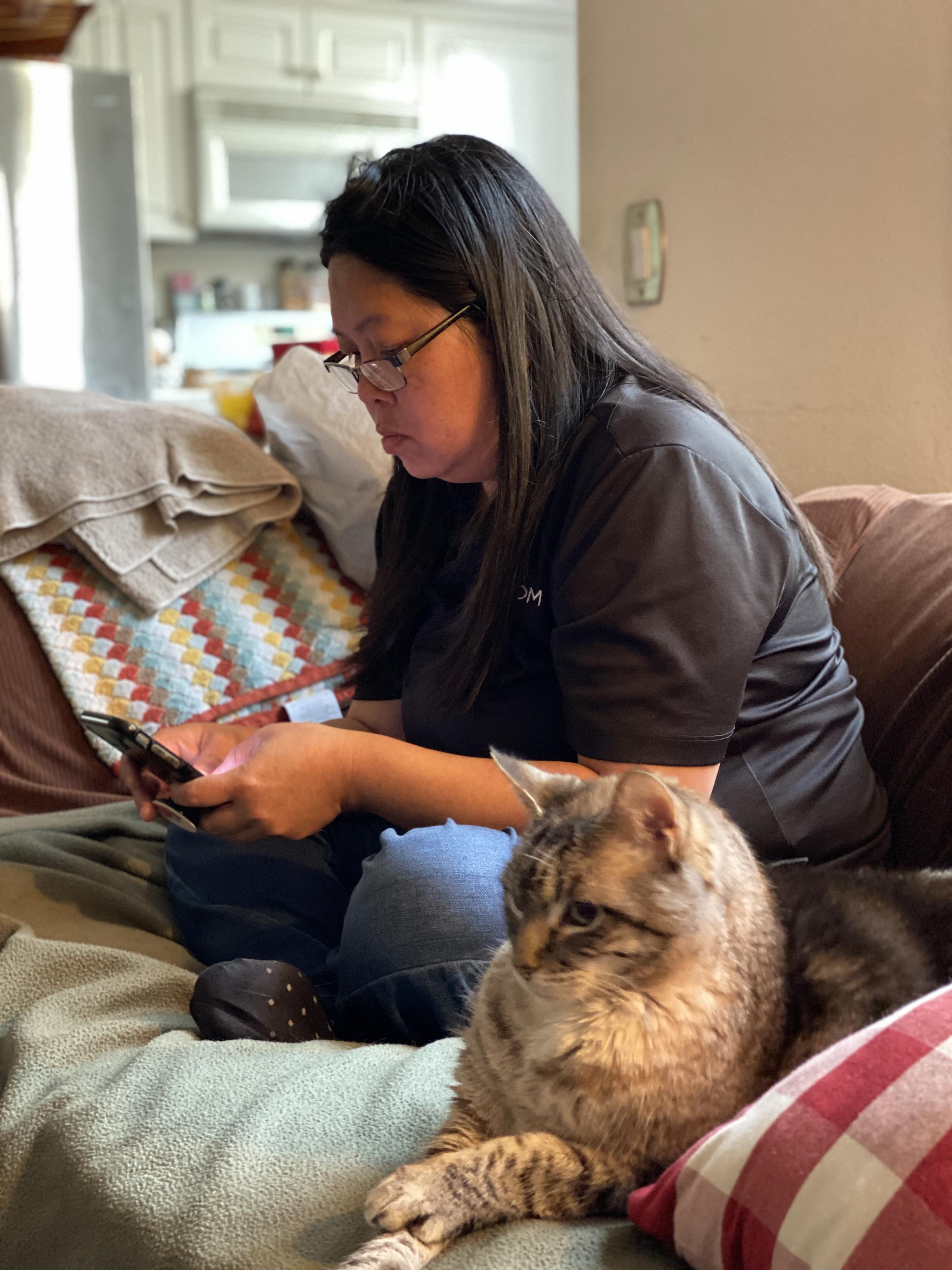 Tam on a couch her legs crossed, looking at her phone, with a Helen lying on a blanket nearby with her paws crossed her head, upright, looking straight ahead.
