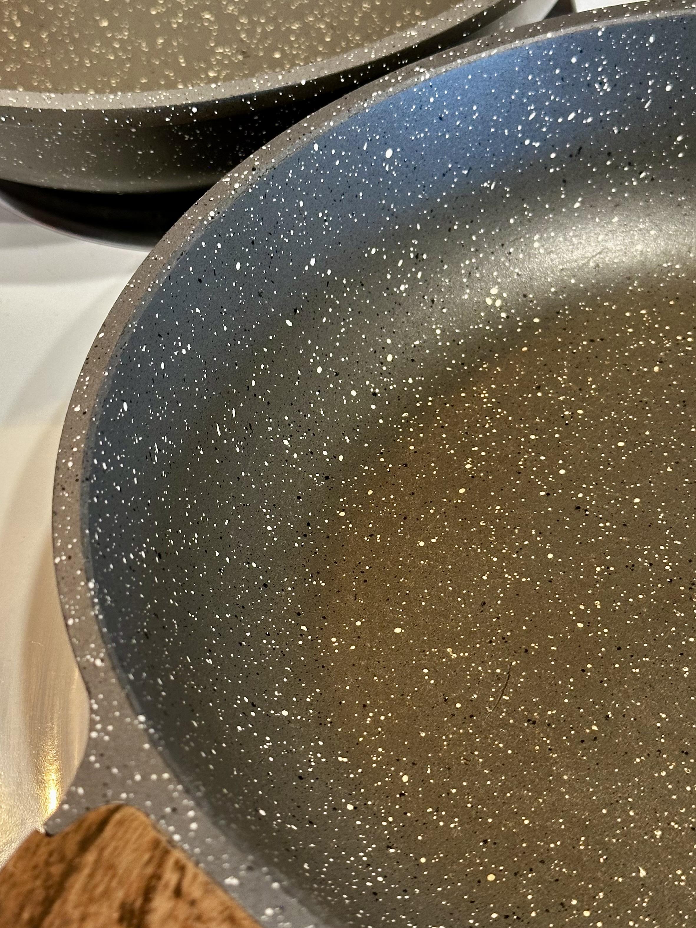 Before vinegar and water stain treatment there is a noticeable brown stain on the inside bottom of each of these medium gray specked skillets that have a ceramic coating to be nonstick. Foreground pan closeup is shown in the After photo next image. They are on a white electric stovetop and have wood-look brown handles.