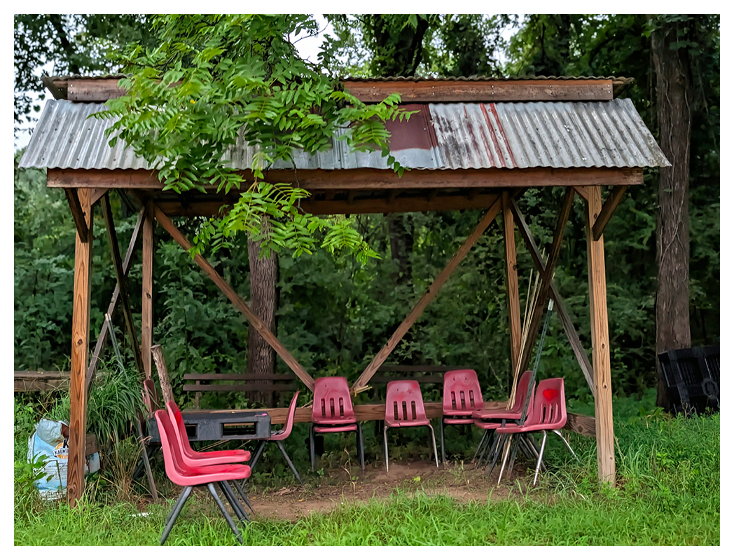 A rustic wooden shelter with a tin roof stands in a grassy clearing. Under it are eight red plastic chairs arranged in a semicircle. Trees and lush greenery fill the background,
