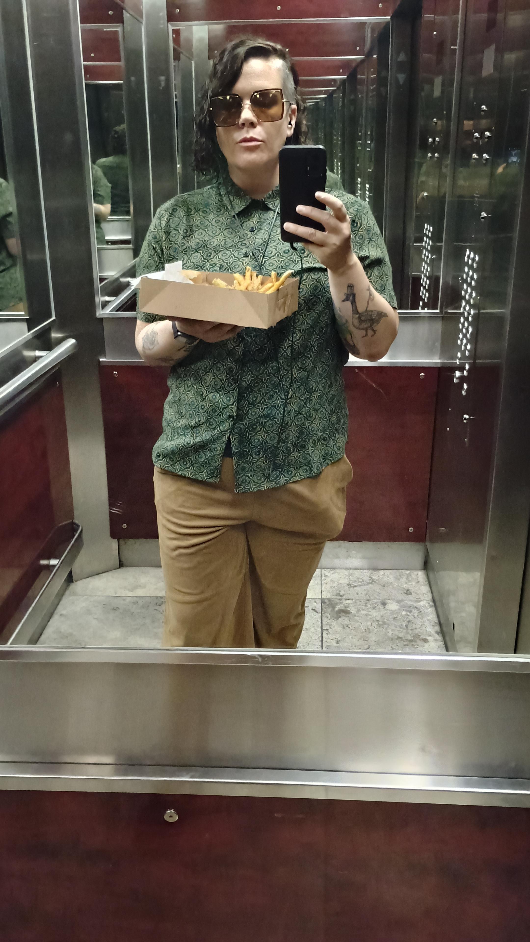 elevator mirror selfish with square sunglasses with brown frames and lenses,  a buttoned up green Hawaiian shirt with tan shapes making a kind of boho pattern, and tan fake suade pants. I am holding a box with fries and a hot dog over my chest and there is a shape visible in my pockets that is either a dick of massive girth or a can of iced tea. probably the dick.