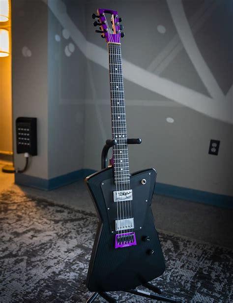 A black guitar with a neck and bridge in a vibrant shade of purple. The guitar is on a stand on a black and white rug. The wall in the background is gray with painted white lines and dots to create a kind of laser light show effect. There is an audio snake attached to the wall.