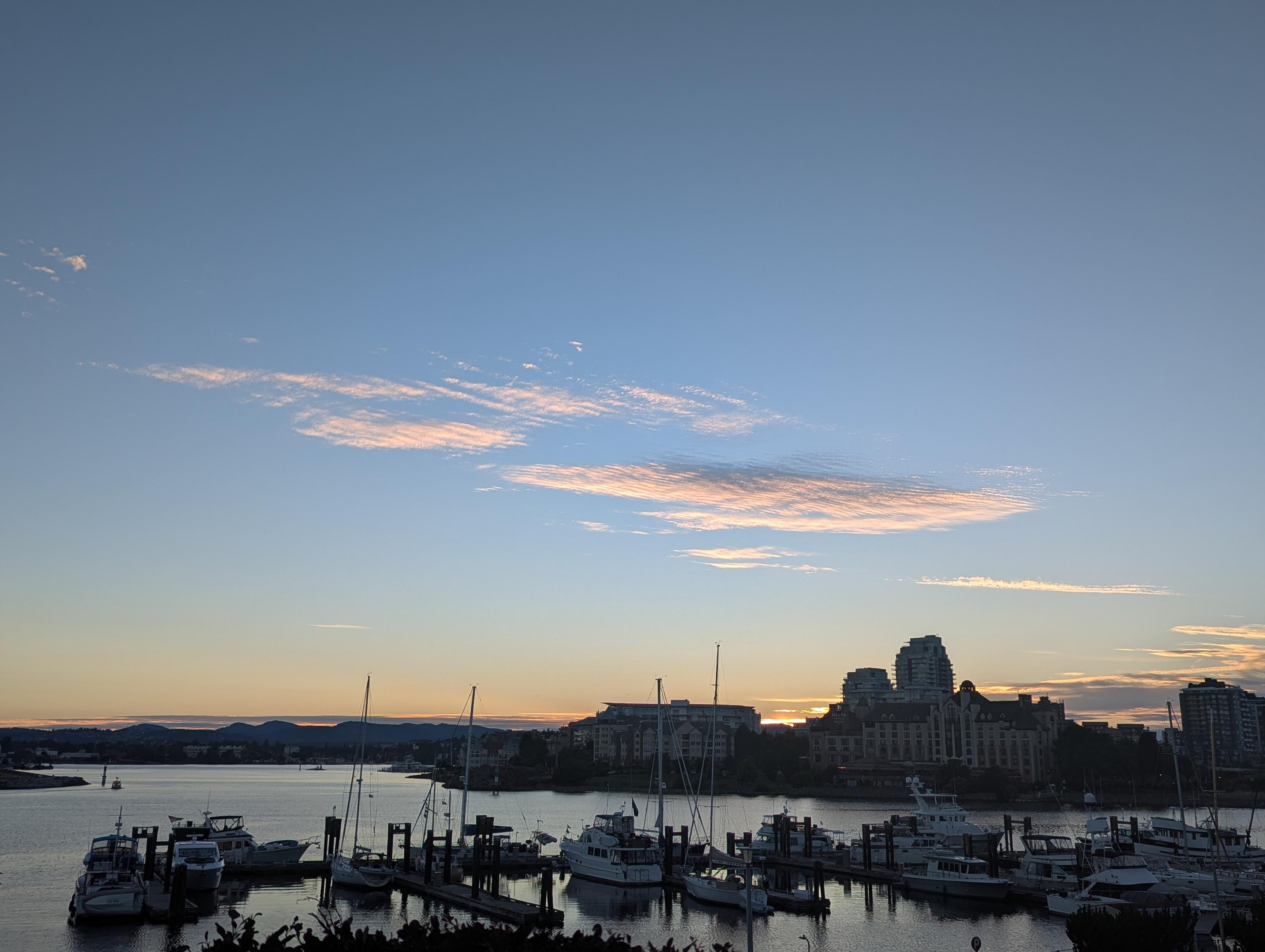 Sunset over Victoria Harbour. The sun is just about set behind the hotel in the distance. You can see the thin band of remaining sun peaking out between the buildings. The sky is a gradient of orange to faded day-blue to dusk-blue. There is a band of clouds lit orange in the middle of the photo.
In the foreground, you can see the boats parked in the harbour.