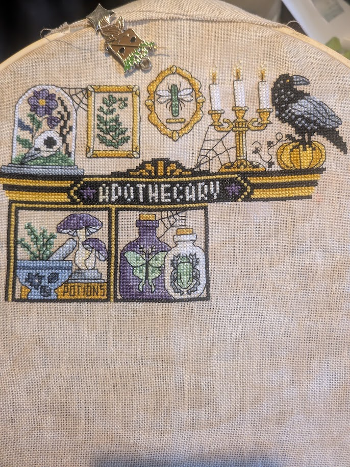 Not So Scary Apothecary cross stitch. 

Its an apothecary cabinet, or will be once its finished.  The cabinet is black and gold, the word apothecary in white at the top of the cabinet.
On top of the cabinet from left to right are - a bell jar with skull and flower inside, gold framed picture of a fern, gold framed picture of a dragonfly, a golden candleabra with 3 white candles and a cobweb reaching from it to the cabinet, finally a raven standing on a pumpkin. 
Below that are the first two spaces in the cabinet, on the left is a pestle and mortar with herbs in it and two purple and white mushrooms, the word potions is also in this space. The next space (which I have just finished) has two bottles one its purple and contains a green moth (or butterfly), the other is white and contains a frog (or toad), both bottles have a cork in them and a cobweb reaches from the cabinet to the top of the bottles.