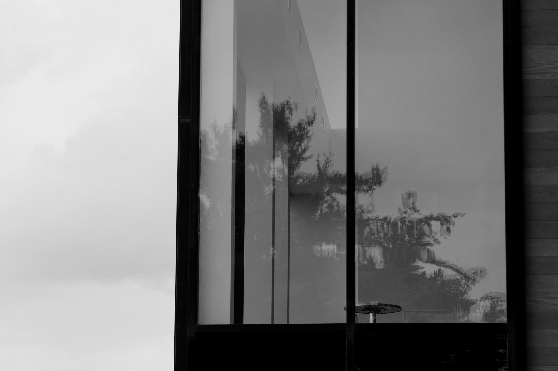 A black and white photograph of a high window, made of two elongated panes of glass with a black frame, on the corner of a building with horizontal wooden slats, visible on the right. To the left, the cloudy sky. The panes reflect the image of tall conifers. Inside, a simple, empty room, except for a small round bistro table.

Photographie noir et blanc d'une fenêtre en hauteur, formée de deux vitres allongées avec un cadre noir, sur le coin d'un bâtiment en lattes de bois horizontales visibles à droite. À gauche, le ciel nuageux. Les vitres reflètent l'image de grands conifères. À l'intérieur, une pièce simple et vide, sauf pour une petite table ronde bistro.