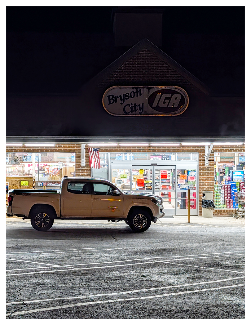 A beige pickup truck is parked outside Bryson City IGA grocery store at night. The store is brightly lit inside, with various items visible through large windows. An American flag hangs near the entrance, and parking lot lines are visible in the foreground.
