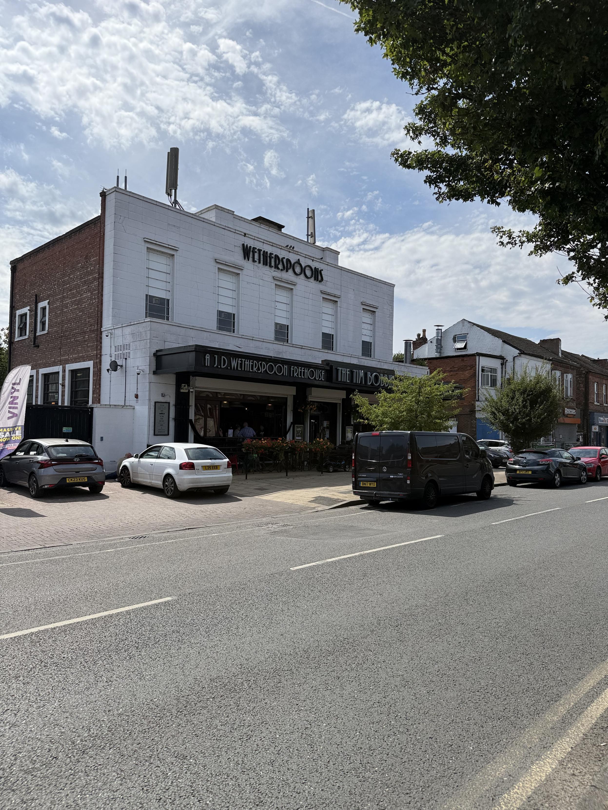 A large square building with a white front stands beside a road. A large black awning above large street levels windows reads “A J D Wetherspoon Freehouse” and just visible “The Tim Bobbin”.  Above the awning are five large windows evenly spaced across the building. Above them in large black lettering is the word “Wetherspoons”
