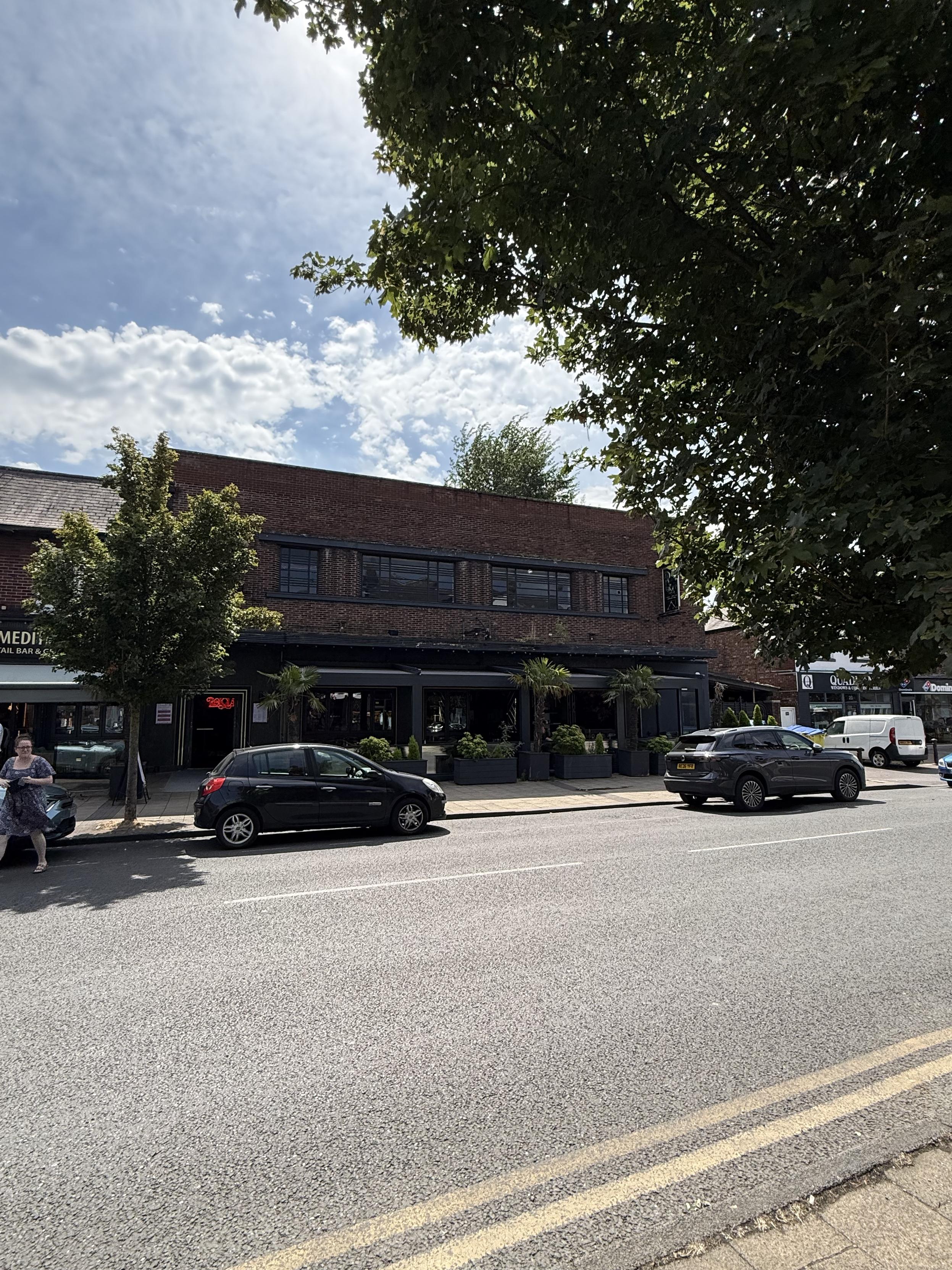 A two or three level dark brick building stands on the side of the road. At street level can be seen a series of interconnecting pergolas in a dark material with tropical planting below them. Some white tables are just visible.  Above can be seen 4 dark framed windows - two large flanked by two smaller ones.