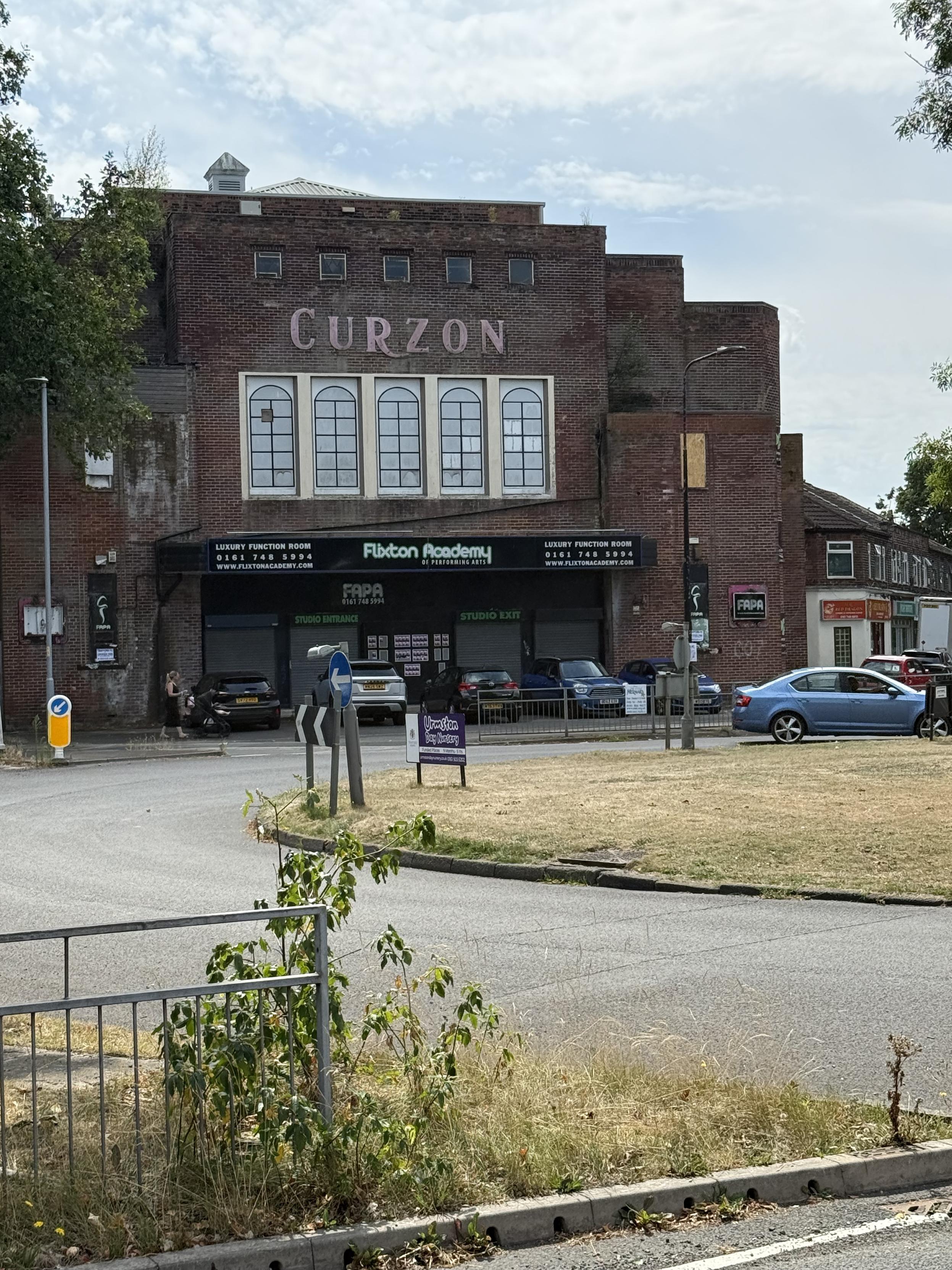 A large brick building stands across a roundabout.  Above a row of 5 large windows it says CURZON. Below is a large black awning/marquee with the words Flixton Academy (and others) on it