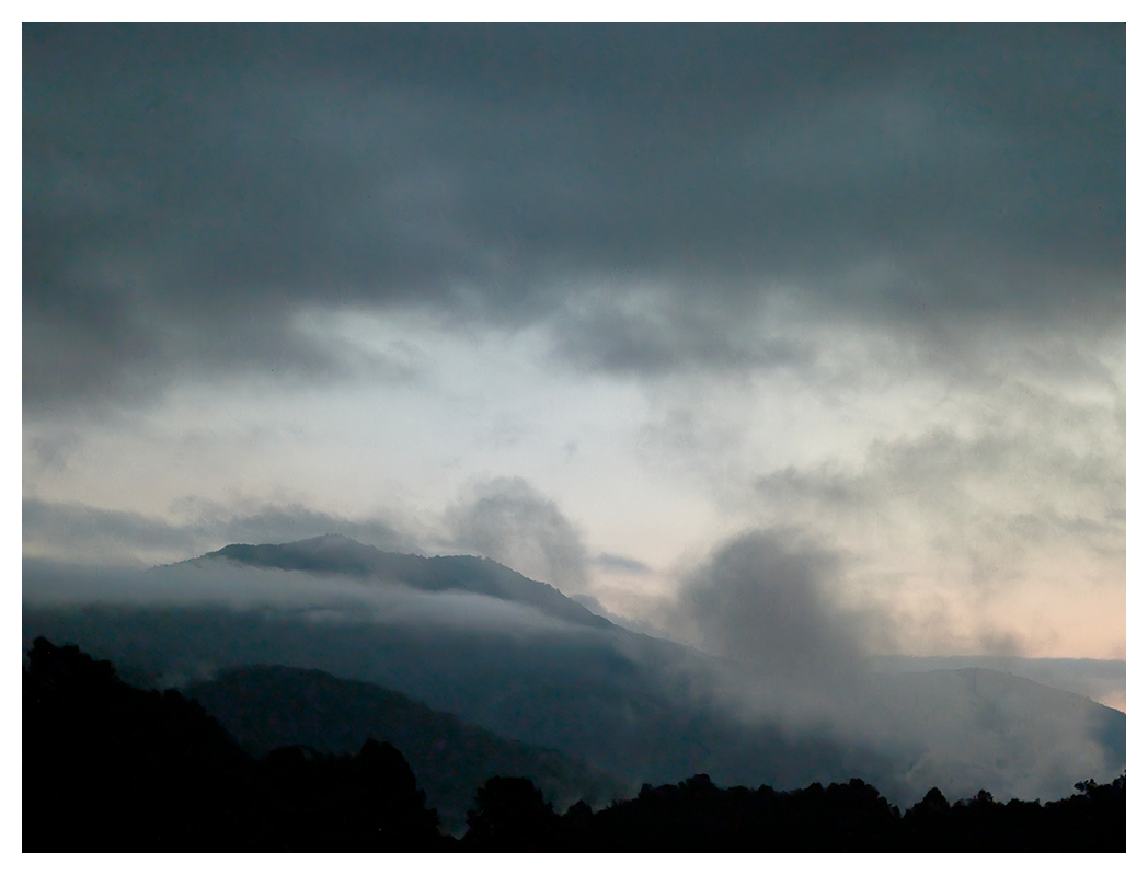 Dark mountains with mist and low clouds fill the foreground, while the sky above is overcast with thick gray clouds. faint light on the horizon, just before sunrise.