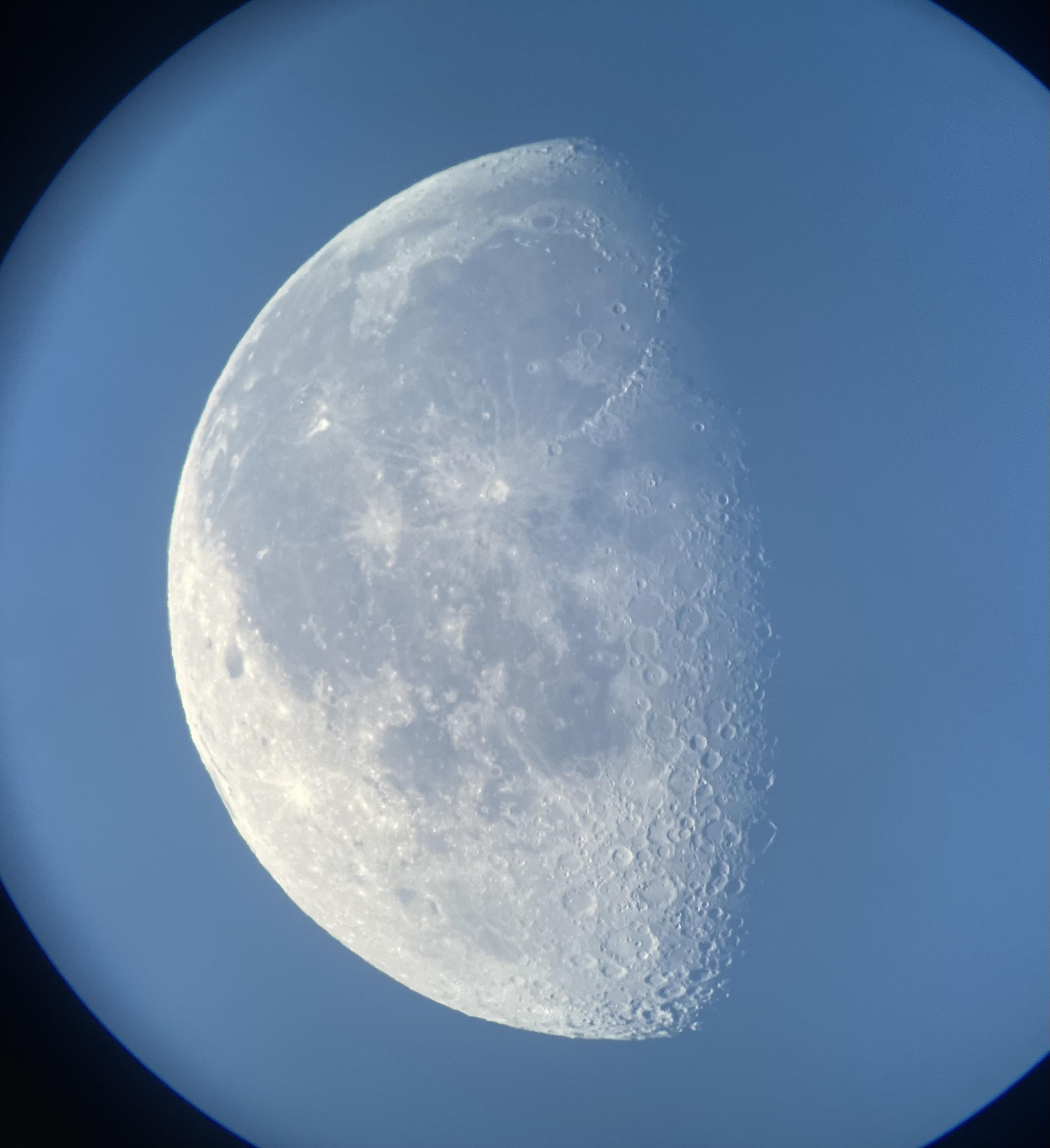 a waning, nearly last quarter moon, pointing to the left in a blue sky. numerous craters can be seen on the terminator lower right, as can darker maria regions on the upper half of the moon