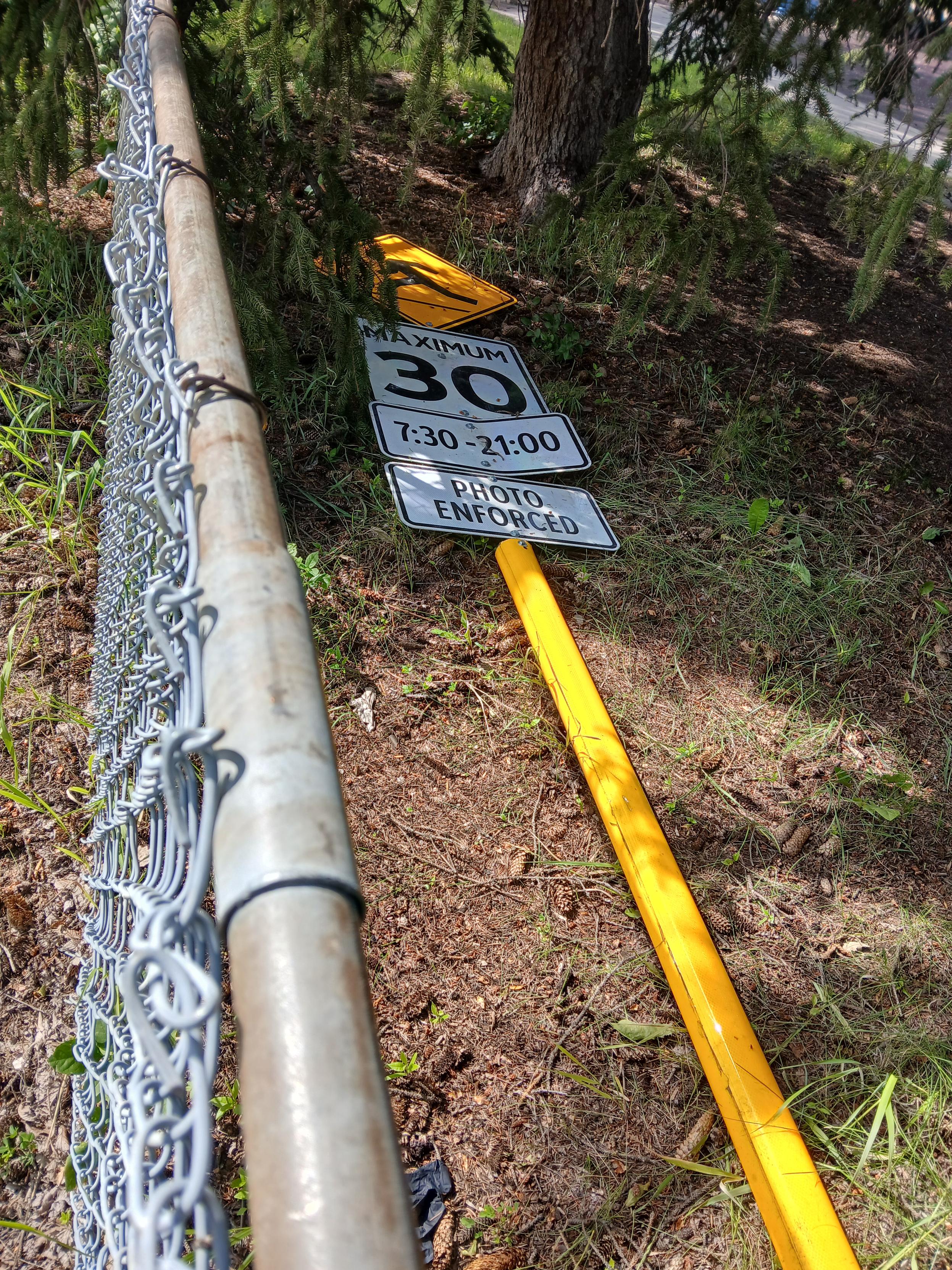 A 30 km/h playground zone speed limit street sign on a high-vis post laying discarded on the inside of a chainlink playground fence in the trees