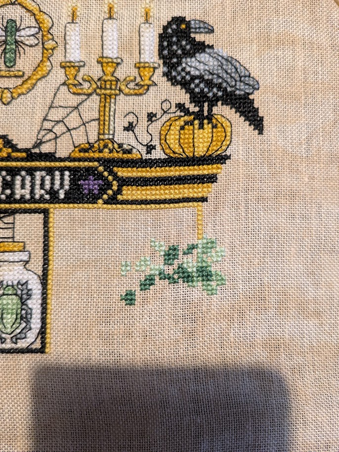 Cross stitch in progress photo, below the raven on the pumpkin another apothecary box is appearing, not much to see so far apart from a cluster of green stitches.