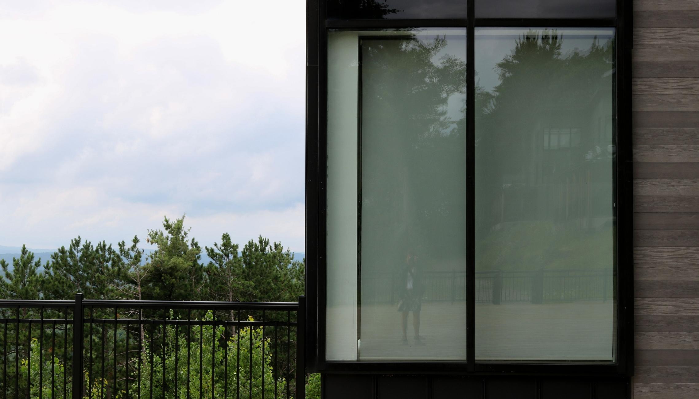 Photograph of a large window framed in black on the corner of a brown slatted building, visible to the right of the window. To the left of the window and the building, a green forest landscape under a cloudy sky stretches beyond a black iron fence. Inside the window, a white canvas has been lowered. Reflected in it is the image of a woman taking the photograph on a wooden patio enclosed by a black fence, with another building and trees behind.

Photographie d'une grande fenêtre entourée d'un large cadre noir sur le coin d'un bâtiment en lattes brunes, visibles à droite de la fenêtre. À gauche de la fenêtre et du bâtiments, un paysage de forêt verte sous un ciel nuageux s'étend de l'autre côté d'une clôture noire en fer. À l'intérieur de la fenêtre, une toile blanche est descendue. L'image d'une femme en train de prendre la photo sur un patio en bois entouré d'une clôture noire s'y reflète, avec un autre bâtiment derrière et des arbres.