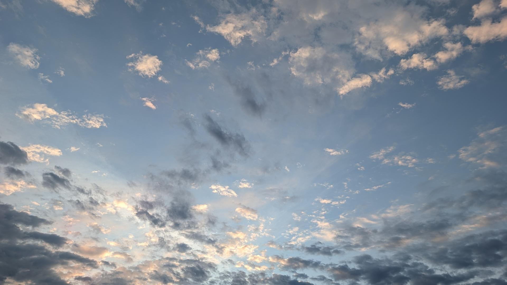 A photo of the early morning sky, a blue gradient backdrop with overlays of clouds. The closest to the ground are a deep grey, some thick with a trail of dancing grey whisps through the center of view. Above those are a layer of white, poofy clouds, reflecting the waking sun. Taken in West Jordan, Utah.