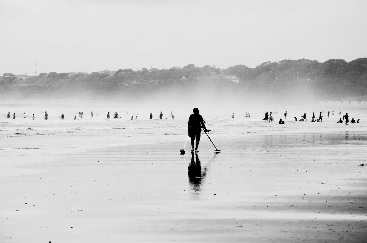 A wide, sandy beach rendered glassy by the retreating tide. A solitary figure mirrored in the surface of the beach sweeps with a metal detector in his right hand. Beyond him, contrasting the misty clouds created by surf, are perhaps a hundred figure doing beachy things, paddling, wading squatting and kneeling in the waves. They are dwarfed by the treasure hunter. And beyond them is a low backdrop comprising trees and the roofs of various buildings.