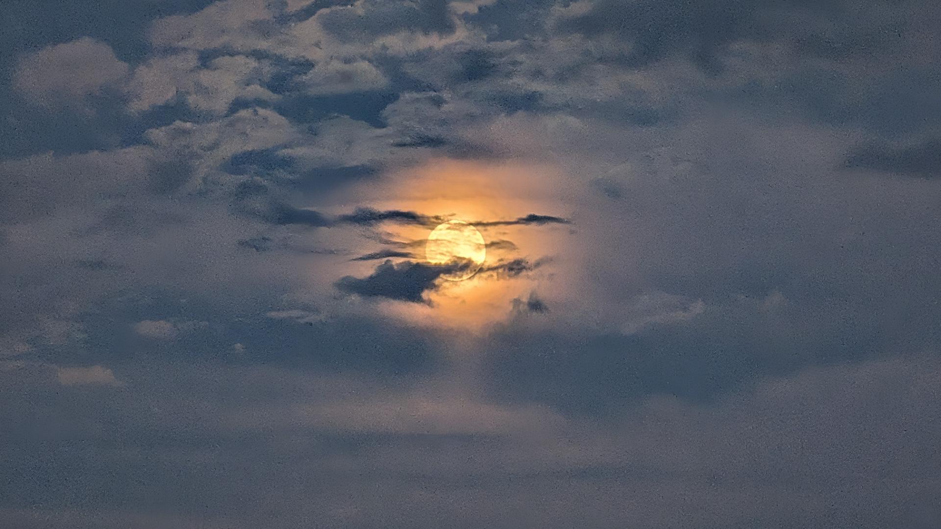 A photo of the moon at sunset. The sky has a baby blue tone from the layer of white clouds, with the full moon's golden glow shining off of the surrounding thin clouds, and a few small, dark whisks layered in front of the moon.
