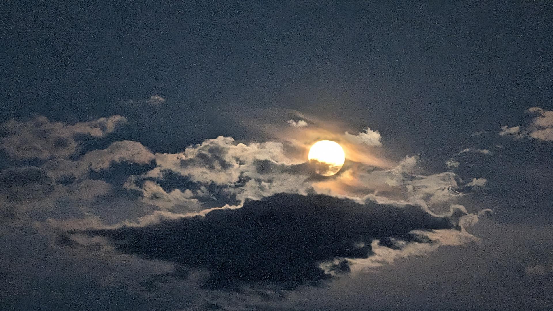 A photo of the moon at dusk. The deep blue sky offsets the puffs of clouds glowing white from the full moon's radiant light. A dense grey cloud floats below the moon, seeming to give the other entities in the sky a platform. Taken in West Jordan, Utah.