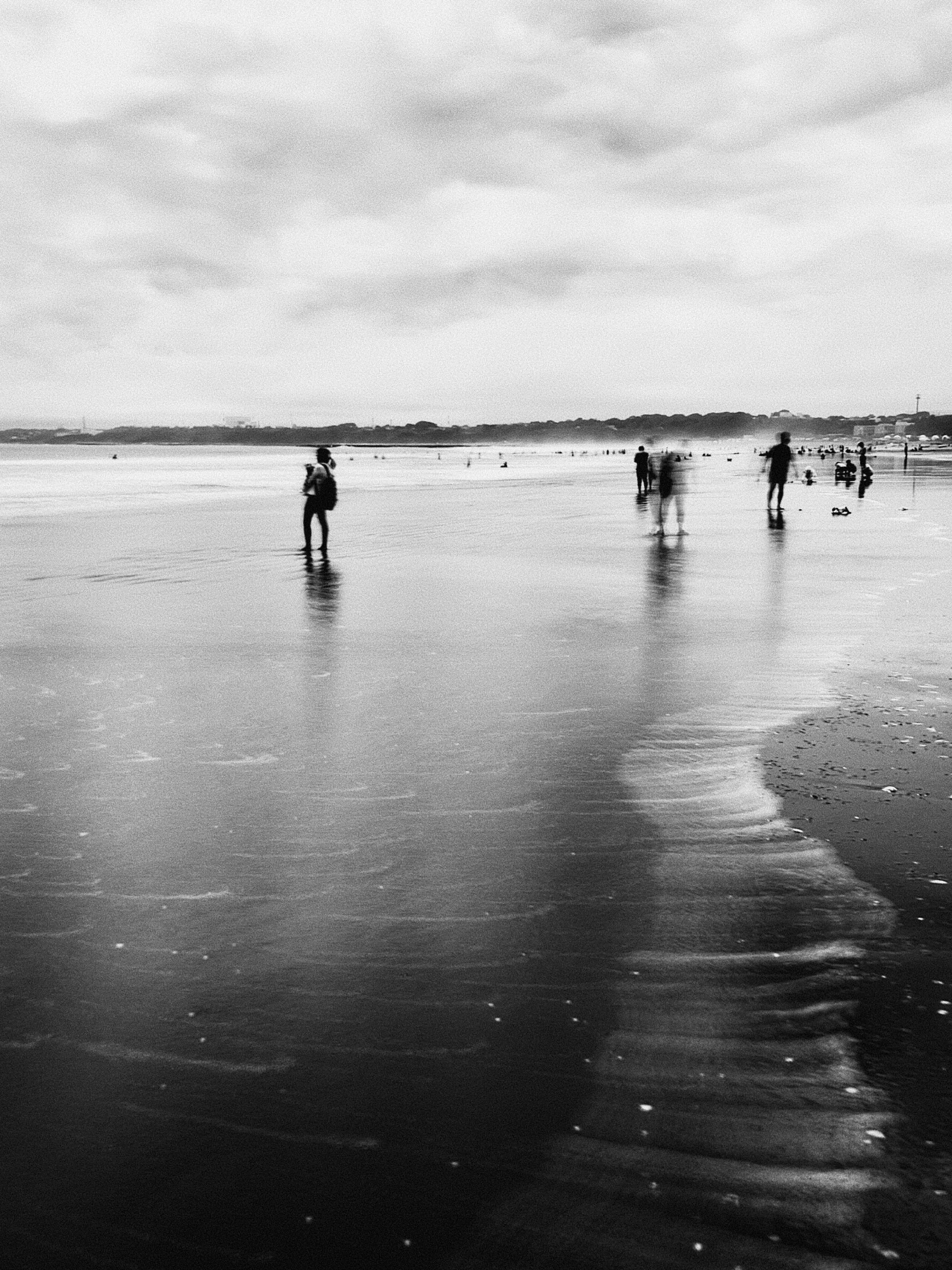 On a wide dark sandy beach the remnants of a wave leave a filament of white surf drifting back to sea. It is scraped like an elongated bar code. At the mid point of this portrait framed shot stand four or five figures pausing in the shallows. They carry bags and buckets, possibly other beach kit but it is difficult to tell because a long expose has both frozen and blurred parts of their profile. Many more beachgoers can be seen in the far distance, blips against the billowing white surf. Beyond every one is a strip of land, trees and some buildings but the contrast renders it a silhouette. The sky is overcast and cloudy.