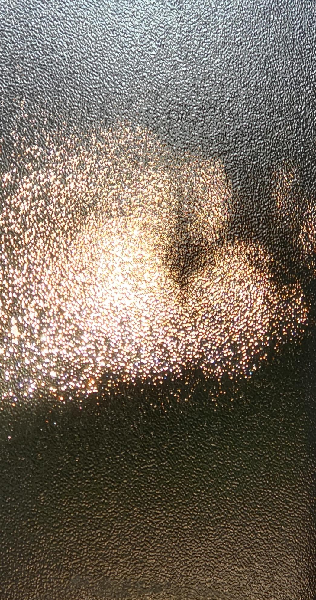 A textured, frosted glass surface with a bright, shimmering pattern of light scattered across the upper left and center portions.