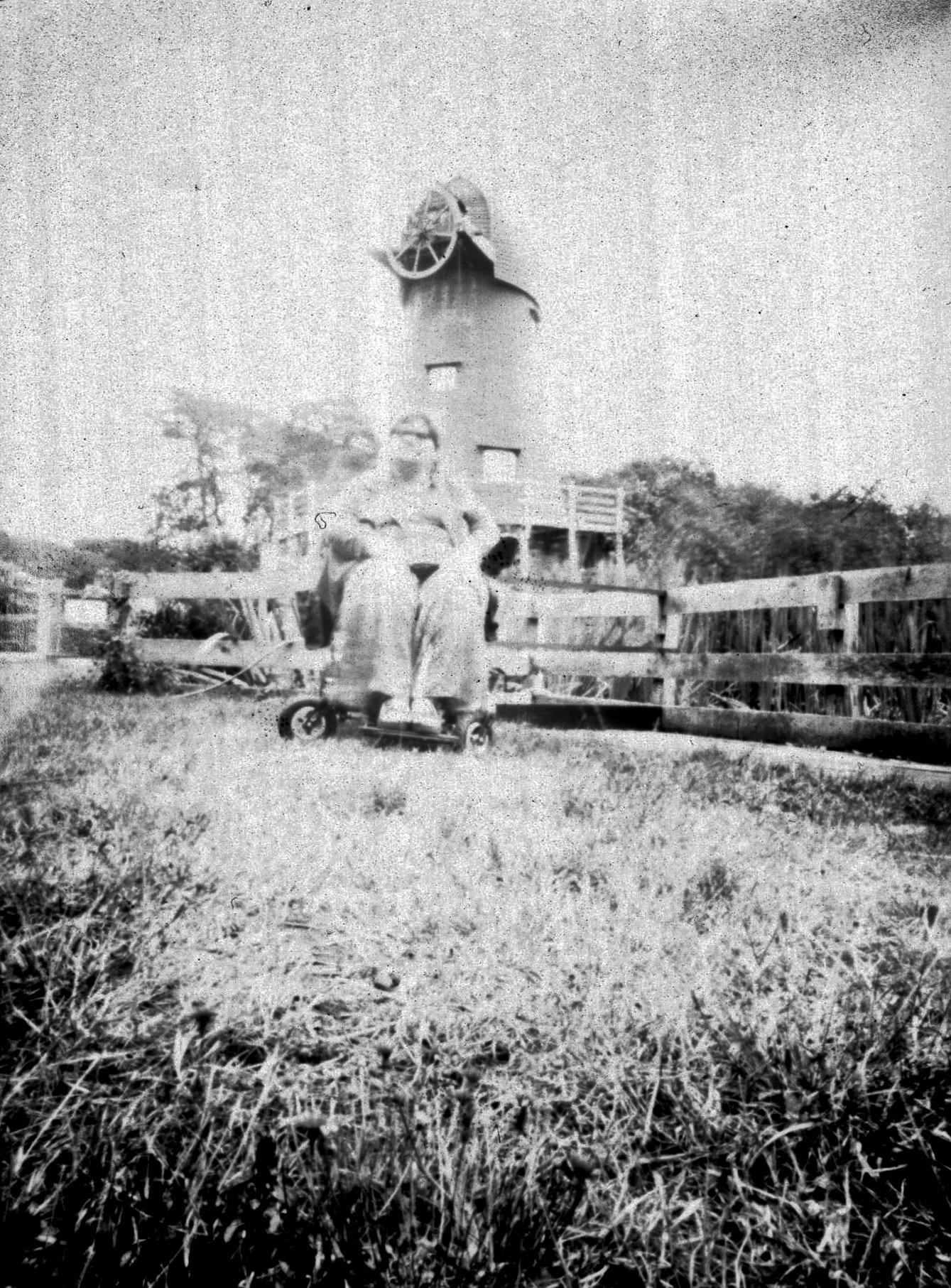 An overexposed black and white shot of me sat in my wheelchair by a fence with an old windmill in the background.