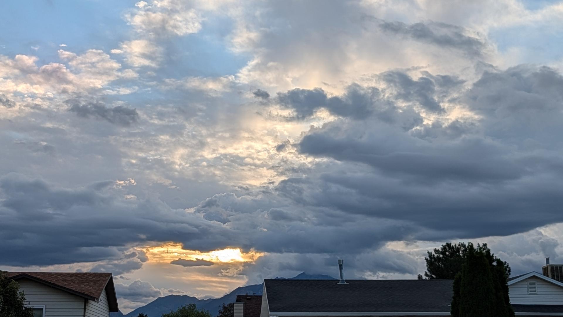 A photo of dramatic clouds, layered over the Salt Lake Valley at sunrise. Behind rooftops are the distant Wasatch Mountains to the east, with layers of large, full clouds, and poofs of others breaking away. A small patch of blue sky is in the upper right corner of this landscape photo, but the clouds take up all the room, with the sun's light showing off in purple hues. A small pale orange glow just above the mountains gives a hint of where the sun lies beyond. Taken from the ground in West Jordan, Utah.