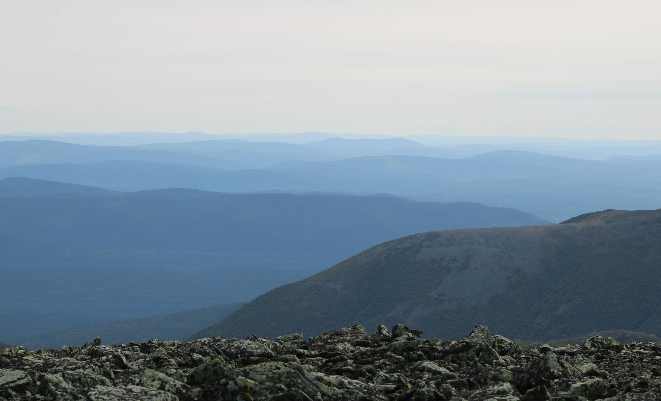 Photograph of a succession of rounded, blue-looking mountains under a very pale blue sky, taken from a higher peak with the rock visible in the foreground below.

Photographie d'une succession de montagnes arrondies et paraissant bleues, sous un ciel bleu très pâle, prise à partir d'un sommet plus haut, dont le roc est visible à l'avant-plan en bas.