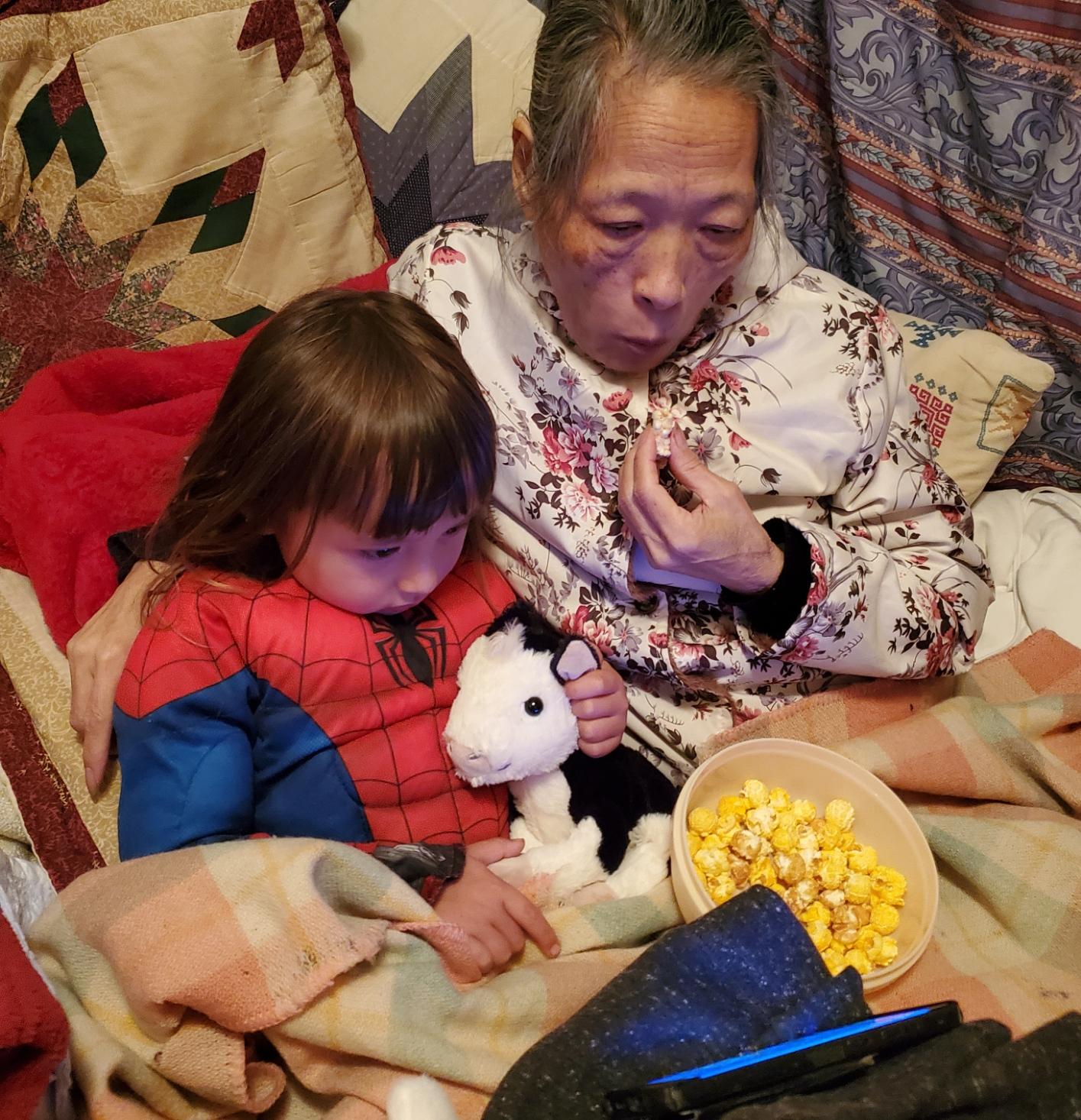 Mom and Rayven (dressed as Spider-Man) sit together wrapped in blankets, watching a movie on a smartphone while eating popcorn from a bowl. He holds a small stuffed cow. It is a cozy and affectionate family moment.